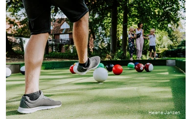 Auf dem Fußballbillardfeld liegen verschieden farbige Fußbälle, die wie große Billardkugeln aussehen. Ein junger Mann schießt dem weißen Ball in Richtung des roten Balls. Seine Frau und seine Kinder (ein Junge und ein Mädchen) schauen ihm beim Spiel zu.