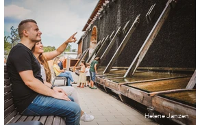 Ein Pärchen sitzt auf einer Bank vor dem Gradierwerk. Sie zeigt auf das Gradierwerk und erzählt ihrem Freund etwas.