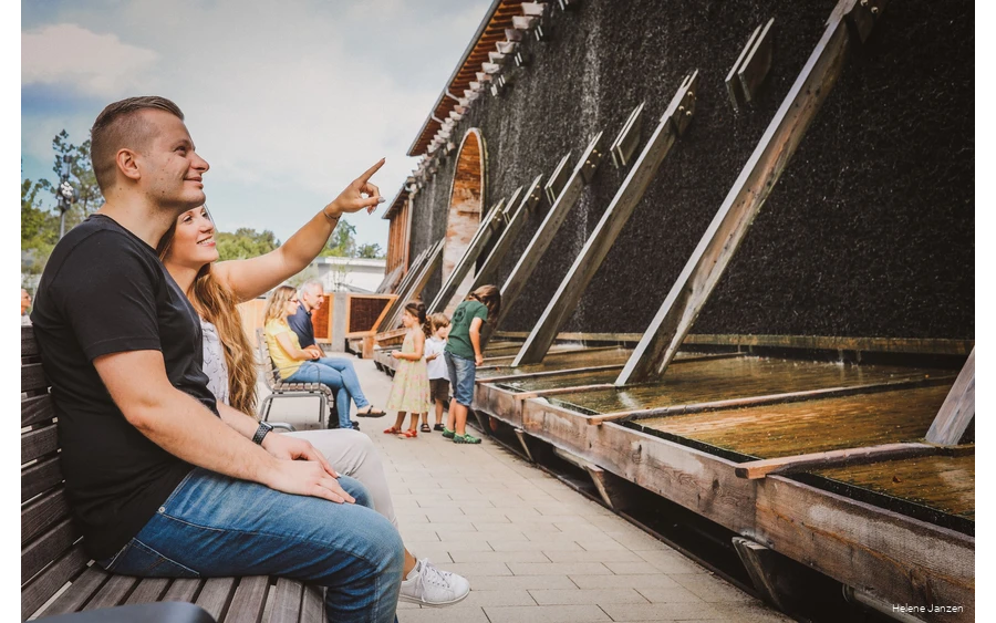 Ein junges Pärchen sitzt auf einer Bank am Gradierwerk im Kurpark Bad Sassendorf. Sie zeigt mit dem Finger in Richtung Gradierwerk und erläutert, warum die Sole am Gradierwerk herunterläuft.