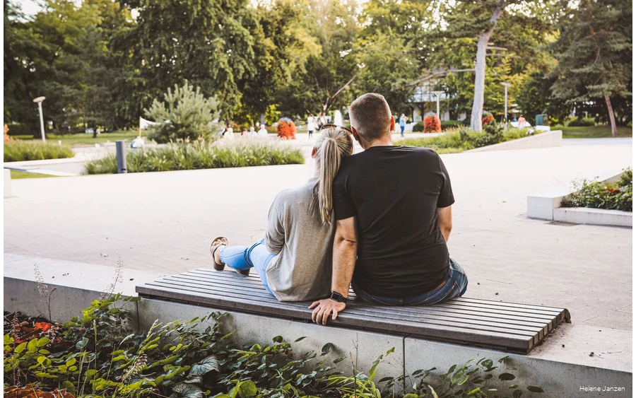 Ein junges Pärchen sitzt auf einer Bank im Kurpark. Drumherum sind schöne Blumenbeete angelegt. Leute spazieren im Kurpark.
